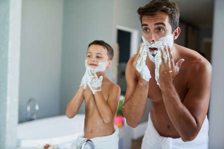 The shaving cream always comes first. Cropped shot of a handsome young man teaching his son how to shave in the bathroom.の写真素材