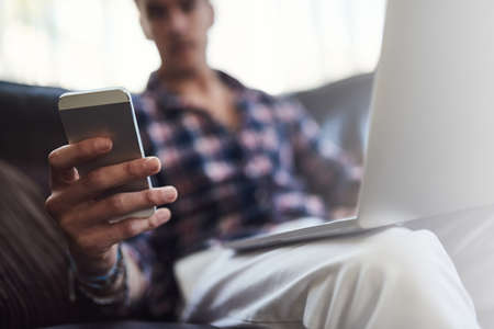 Social networking on multiple platforms. Shot of a young man using his laptop and cellphone while sitting on the sofa at home.の写真素材