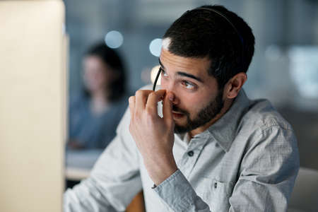 Just when you thought you could clock out. Shot of a young man feeling stressed while using a headset and computer late at night in a modern office.の写真素材