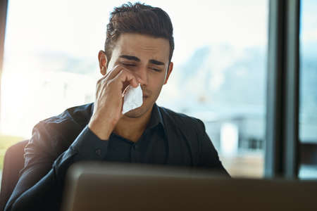 Feeling a cold coming on. Shot of a young businessman blowing his nose in an office.の写真素材