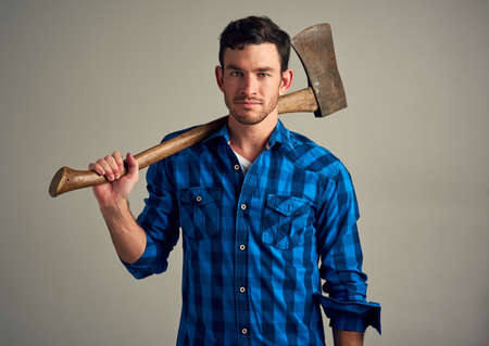 Need something chopped down. Studio shot of a young male lumberjack holding an axe against a grey background.の写真素材