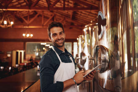 Im always happy when Im at work. Portrait of a cheerful young barman browsing on a digital tablet while keeping track of stock inside of a beer brewery during the day.の写真素材