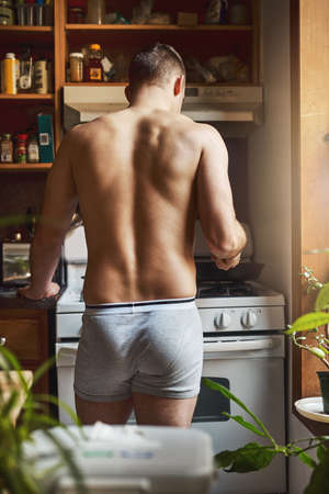 Lets get cooking then eating. Rearview shot of an unrecognizable shirtless man cooking in the kitchen at home.の写真素材
