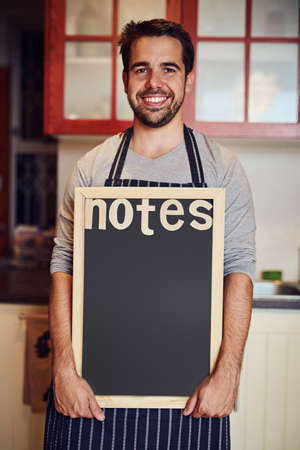 Make a note of it. Portrait of a happy young man holding a blank chalkboard while standing in his kitchen at home.の写真素材