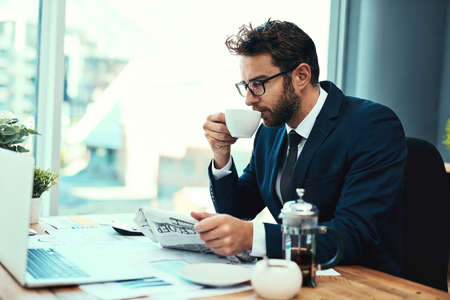 Lets see how the markets are doing. Shot of a young businessman drinking a cup of tea while reading a newspaper in an office.の写真素材
