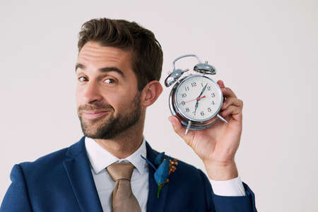 The wait is over. Studio shot of a handsome young groom holding a clock against a gray background.
