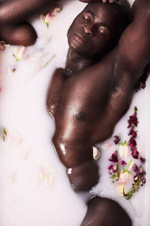 You cant beat the relaxing power of a bath. Shot of a muscular young man having a milky bath filled with flowers at home.の写真素材