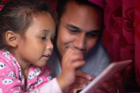 Story time is her favorite part of going to bed. Shot of a father reading a story to his little girl from a digital tablet while lying under together under a blanket.の写真素材