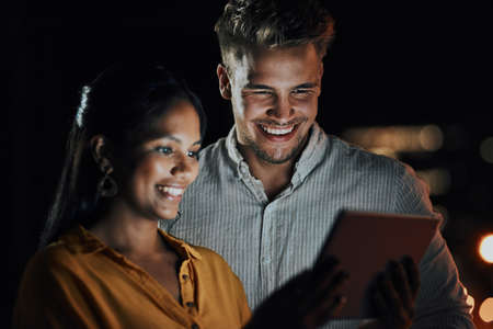 This app is perfect for what we need. Shot of two businesspeople using a digital tablet together outside an office at night.の写真素材