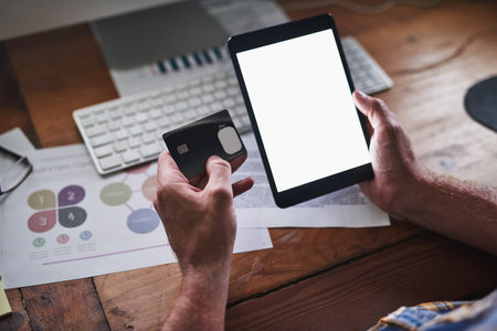 Well deliver right to your desk. Shot of an unidentifiable programmer holding his credit card while using his tablet in the office.の写真素材