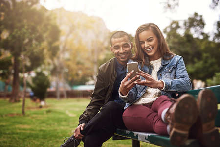 What do you think about this photo. Shot of a cheerful young couple sitting down on a bench while using a phone together outside in a park.の写真素材