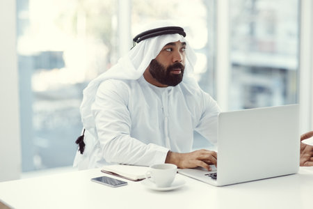 Hes got an idea. Cropped shot of a young businessman dressed in Islamic traditional clothing working on his laptop while sitting in the office.の写真素材