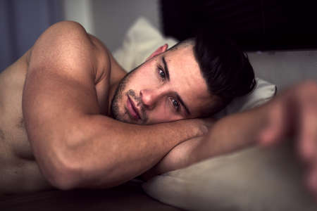 My bed, or yours. Portrait of a shirtless young man giving an inviting look while lying in bed.の写真素材