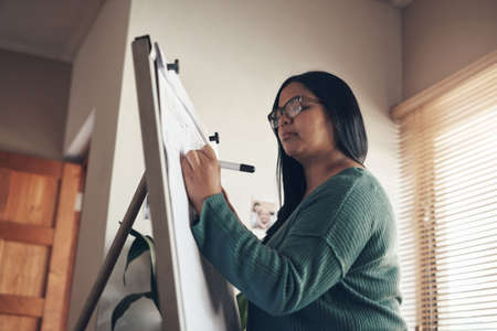 Class in quarantine is now in session. Shot of a young woman using a whiteboard to teach a lesson from home.の写真素材