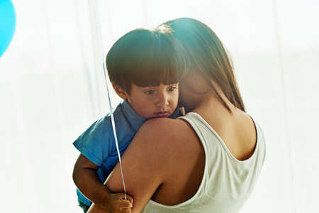 Safe in moms embrace. Shot of a young mother cuddling her little boy while he holds a balloon at home.の写真素材
