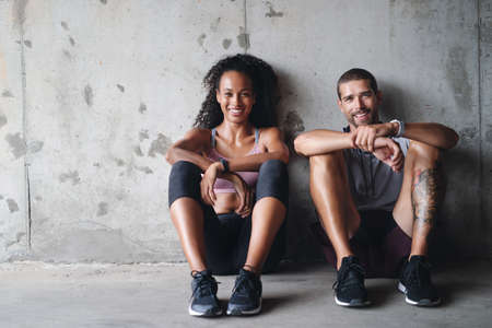 Were down but not out. Portrait of a sporty young couple sitting down against a wall while exercising inside a parking lot.の写真素材