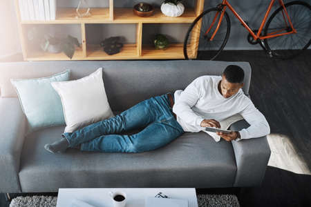 I cant manage to find the app I just downloaded. High angle shot of a young man lying down on a couch and using his digital tablet at home.の写真素材