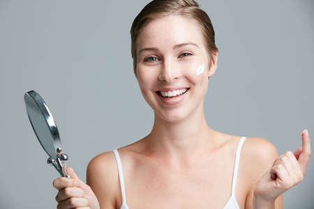 Be kind to your skin. Youve got it for life. Studio portrait of an attractive young woman holding a hand mirror and applying cosmetics against a gray background.の写真素材