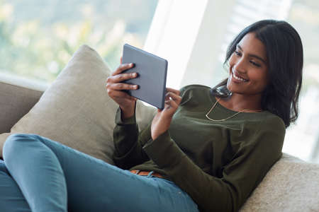 Checking her social media feeds. Shot of an attractive young woman using her tablet while sitting on the sofa at home.の写真素材