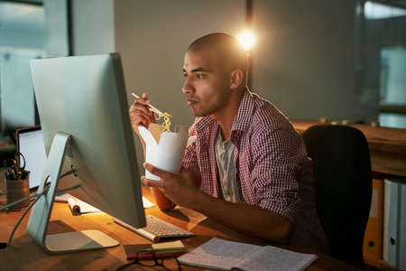 Success is the order of the day and night. Cropped shot of a young designer eating takeaways while working late in an office.の写真素材