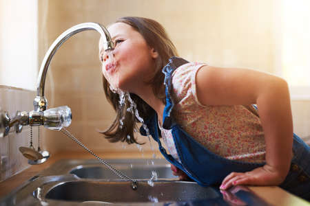 The thirst is real. Shot of a little girl drinking water directly from the kitchen tap at home.の写真素材