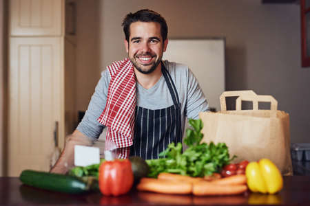 Cooking well doesnt mean cooking fancy. Portrait of a happy young man posing behind a selection of fresh ingredients in his kitchen.の写真素材