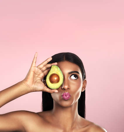Improving her skin, one avocado at a time. Studio shot of a beautiful young woman posing with an avocado against a pink background.の写真素材