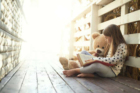 Which story should we ready next teddy. Shot of a little girl reading a book with her teddy bear beside her.の写真素材