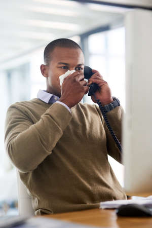 Making an appointment to see his gp. Shot of a young businessman blowing his nose at his work desk.の写真素材