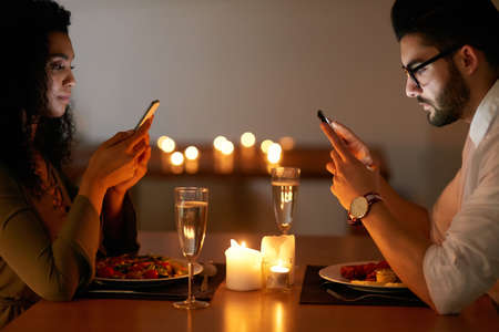 Distracted over dinner. Shot of a young couple texting on their phones while out for dinner.の写真素材
