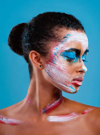Always put your best side forward. Studio shot of a beautiful young woman covered in face paint posing against a blue background.の写真素材