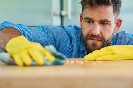 Powering through household chores like a champ. Shot of a man wearing rubber gloves while wiping a table at home.の写真素材