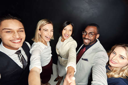 Who said we couldnt take selfies at work. Portrait of a group of businesspeople taking a selfie against a dark background.の写真素材