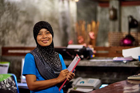 Youre always welcome here. Portrait of a smiling muslim thai woman standing behind a hotel reception desk holding a clipboard.の写真素材