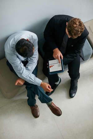 Going over the daily numbers. High angle shot of two businessmen talking together over a digital tablet while sitting in a modern office.の写真素材