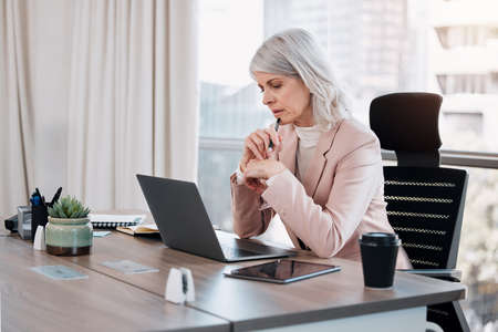 Hmmm, I need to think. Shot of a mature businesswoman sitting alone in the office and looking contemplative while using her laptop.の写真素材