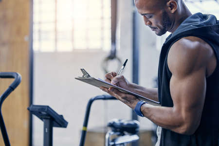 Organising and stocking supplies for all gym patrons. Shot of a muscular young man writing notes on a clipboard while working in a gym.の写真素材