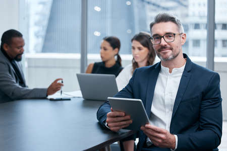 Were always busy making big things happen here. Portrait of a mature businessman using a digital tablet in an office with his colleagues in the background.の写真素材