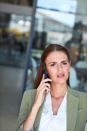 I dont understand...can you explain that again. Shot of a young businesswoman using a mobile phone and looking annoyed in a modern office.の写真素材