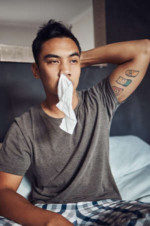 Has anybody seen my tissue. Shot of a young man blowing his nose while recovering from an illness in bed at home.の写真素材