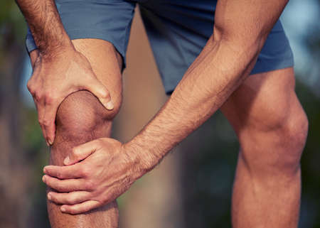 I cant run with this sore knee. Closeup shot of an unrecognisable man holding his knee in pain while exercising outdoors.の写真素材