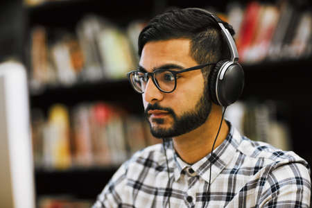 This all going to pay off soon. Closeup shot of a focused young man sitting and working on a computer in a library while listening to music.の写真素材