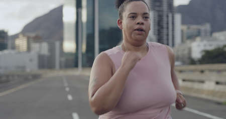 Close up of mixed race woman city running along highway in outdoor workout. Plus size woman starting...