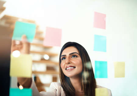 She turns good ideas into great ideas. Shot of a businesswoman arranging sticky notes on a glass...