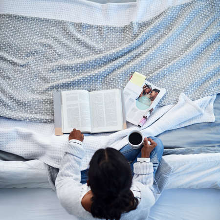 Relaxing with a good read. High angle shot of an unrecognizable woman reading a book and drinking coffee on her bed in her bedroom at home.の写真素材