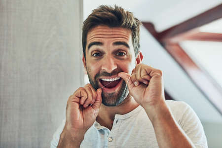 My teeth makes a great smile. Portrait of a cheerful young man flossing his teeth while looking at his reflection in the mirror at home.の写真素材