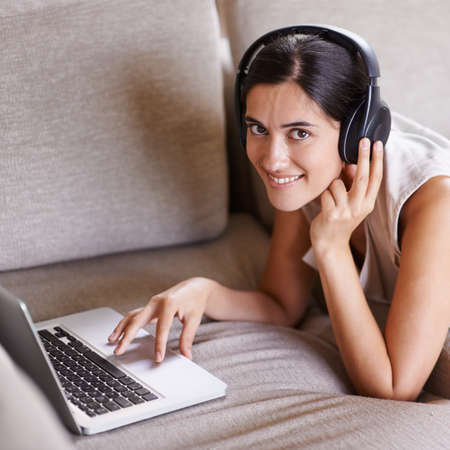 Staying in touch in the digital age. Shot of a young woman on a sofa wearing headphones and using a laptop.の写真素材
