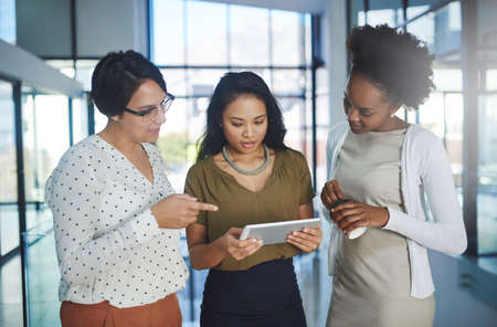 Putting their heads together. Cropped shot of three businesswomen looking at a tablet in the office.の写真素材