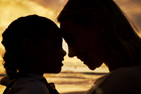 Closeup silhouette of a mother and daughter standing face to face on the beach at sunset. Backlit young woman and her girl child smiling and facing one another with the ocean in the backgroundの写真素材