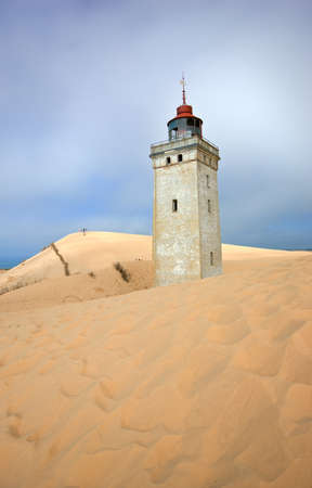 Lighthouse on a sand dune by the sea against a blue sky. Mysterious old tower alone in the desert. Deserted lighthouse isolated on beige sand. Peaceful and tranquil nature scene in Jutland, Denmark.の写真素材